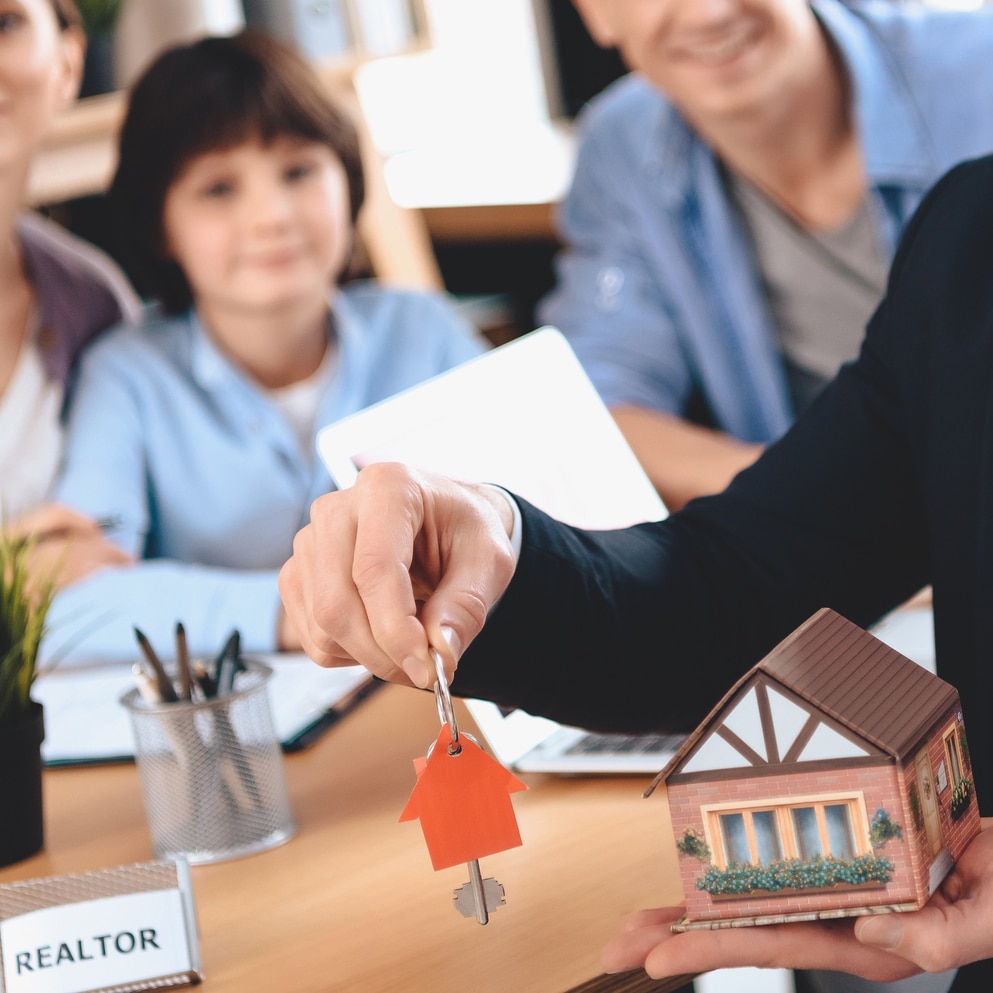 Realtor Sitting At Desk In Office. Realtor Is Presenting Keys And Model House With Family In Background.